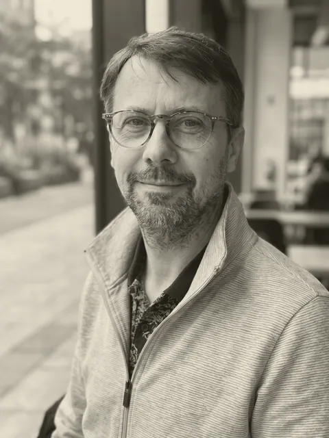 Portrait en noir et blanc de Jérôme Brigaud : lunettes, barbe courte, veste zippée à fines rayures, sourire